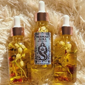 May include: Three glass bottles of Mermaid Aura Ritual Oil with white droppers and copper-colored collars. The bottles contain a golden liquid with floral elements. Each bottle has a label with a mermaid illustration and the text "Mermaid Aura Ritual Oil" and "River Rose Botanica".