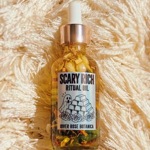 May include: A clear glass dropper bottle filled with golden oil and botanical pieces. The label reads SCARY RICH RITUAL OIL, RIVER ROSE BOTANICA, and features a graphic of a ghost next to a stack of gold bars and roses in black ink.