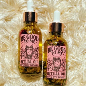 May include: Two glass bottles of ritual oil with gold-colored tops and white droppers. Each bottle has a pink label with the text "BE GOOD TO ME" and "RITUAL OIL RIVER ROSE BOTANICA". The bottles are filled with a yellow liquid and small plant pieces.