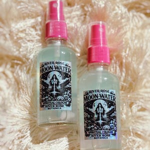 May include: Two clear glass spray bottles with pink spray tops. Each bottle has a label with the text "RIVER ROSE MOON-WATER SPIRITUAL COLOGNE RIVER ROSE BOTANICA" and a graphic of a woman. The bottles are on a cream-coloured, textured surface.
