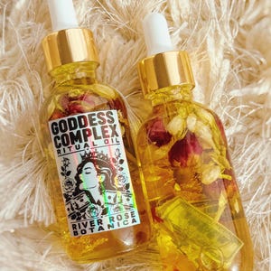 K&ouml;nnte beinhalten: Zwei durchsichtige Glasflaschen mit Pipette, gef&uuml;llt mit gelbem &Ouml;l, getrockneten Rosenknospen und Miniatur-Geldscheinen. Die linke Flasche hat ein wei&szlig;es Etikett mit dem Text Goddess Complex Ritual Oil River Rose Botanica und einer Grafik.