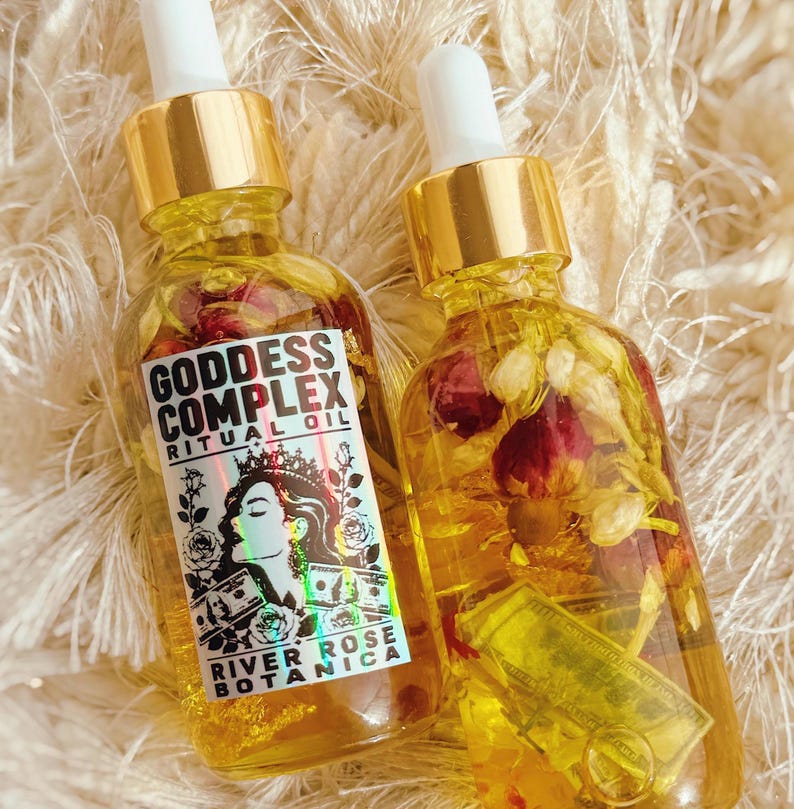 K&ouml;nnte beinhalten: Zwei durchsichtige Glasflaschen mit Pipette, gef&uuml;llt mit gelbem &Ouml;l, getrockneten Rosenknospen und Miniatur-Geldscheinen. Die linke Flasche hat ein wei&szlig;es Etikett mit dem Text Goddess Complex Ritual Oil River Rose Botanica und einer Grafik.