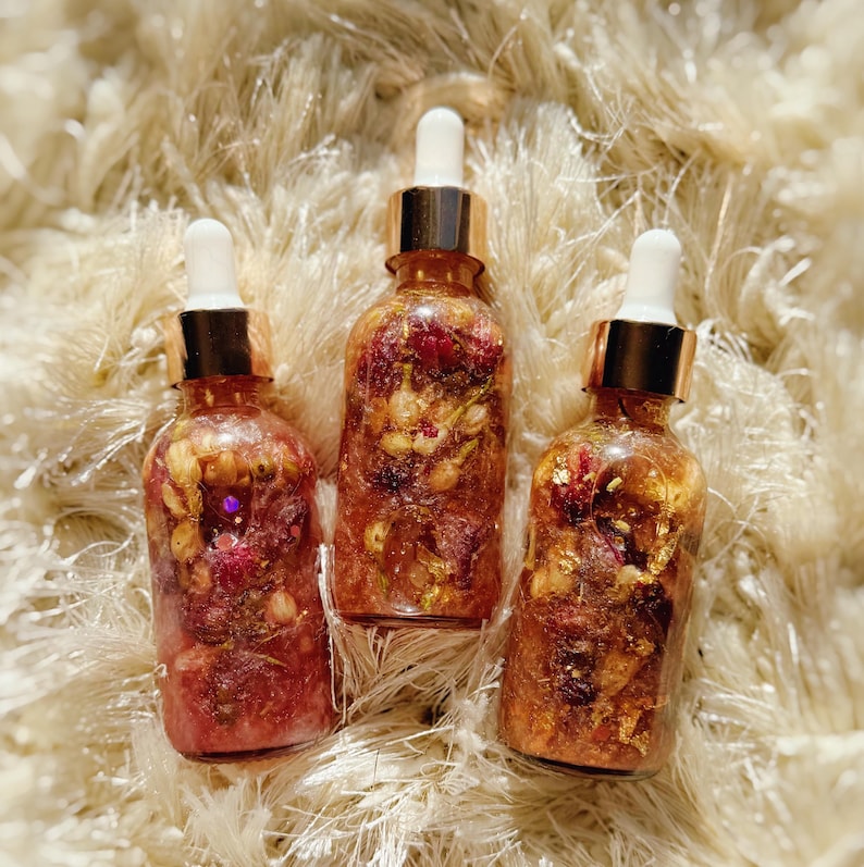 May include: Three glass dropper bottles filled with a clear, golden liquid and floral inclusions. The bottles have gold-coloured caps and white droppers. The background is a fluffy, cream-coloured surface.