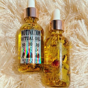 May include: Two clear glass dropper bottles filled with golden oil and botanical herbs. The left bottle features a label with the text MOTIVATION RITUAL OIL 10 10 10 RIVER ROSE BOTANICA. The right bottle contains visible dried leaves and petals.