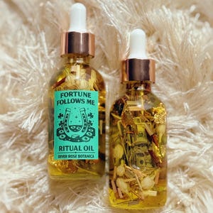 May include: Two glass ritual oil bottles with gold-colored tops and white droppers. One bottle has a teal label with the text "Fortune Follows Me" and a horseshoe graphic. The other bottle is filled with herbs and gold flakes.