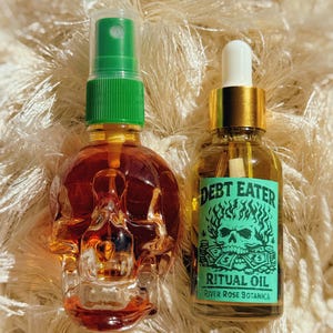 May include: Two glass bottles of ritual oil. One is skull-shaped with a green spray top. The other is a small glass bottle with a gold cap and a white dropper. The label reads "DEBT EATER RITUAL OIL" with a skull and flames graphic.