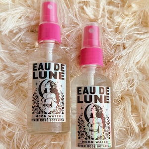 May include: Two clear glass spray bottles with bright pink caps and nozzles. Each bottle features a rectangular label with the text EAU DE LUNE, MOON WATER, and RIVER ROSE BOTANICA, accompanied by a black and white illustration of a person.