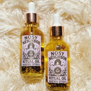 May include: Two glass bottles of ritual oil with dropper tops. The labels read "Nosy Neighbor Ritual Oil River Rose Botanica" with an eye and floral design. The oil is yellow with small particles inside.