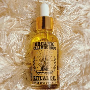 May include: A clear glass dropper bottle filled with golden liquid. The label is tan with black text reading ORGANIC CALAMUS ROOT RITUAL OIL RIVER ROSE BOTANICA and features a black line drawing of a plant with roots and small star accents.