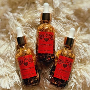 Könnte beinhalten: Drei Glasflaschen mit Ritualöl mit goldfarbener Flüssigkeit und einer Pipette. Jede Flasche hat ein rotes Etikett mit dem Wort "COMMITMENT" und einem Herzdesign. Die Flaschen stehen auf einer flauschigen, weißen Oberfläche.