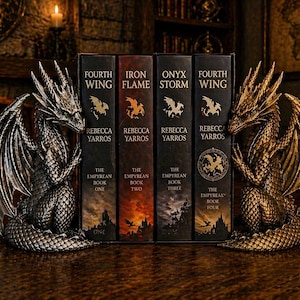Op de afbeelding: Drakenboekensteunen flankeren een verzameling fantasyromans. De boekensteunen zijn gedetailleerde drakensculpturen in zilver- en goudtinten. De boektitels zijn onder meer "Fourth Wing", "Iron Flame" en "Onyx Storm" van Rebecca Yarros.