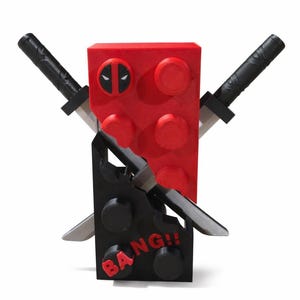 May include: A red and black toy block set with a Deadpool theme. The red block features the Deadpool logo, with two black katanas crossed in front. A black block at the bottom reads "BANG!!".