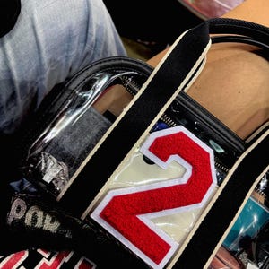 May include: A clear tote bag with black straps and a white stripe. The bag features a large red number "2" patch with a white border. The bag is filled with various items, and the word "Pow" is visible on the side.