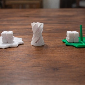 Puede incluir: Tres objetos impresos en 3D sobre una superficie de madera. Una torre blanca retorcida se encuentra entre otros dos objetos. Uno es un cubo blanco sobre una base blanca en forma de estrella con una varilla blanca. El otro es un cubo blanco sobre una base verde en forma de estrella con una varilla verde.
