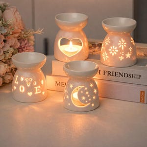 Puede incluir: Cuatro quemadores de aceite de cerámica blanquecina con diseños recortados. Uno tiene un corazón, otro flores, un tercero la palabra "LOVE" y el último una luna y estrellas. Cada quemador tiene un cuenco pequeño y una vela encendida.