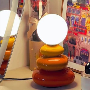 Puede incluir: Una lámpara de mesa decorativa con un diseño de piedras apiladas. La lámpara presenta una luz de globo blanca brillante sobre una base de piedras de cerámica naranja, amarilla y rojo anaranjado. La lámpara está enchufada.