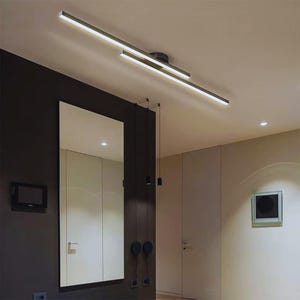 Puede incluir: Luminaria de techo moderna y lineal con dos barras de luz rectangulares paralelas. La luminaria está montada en un techo blanco, con una pared oscura y un gran espejo en el fondo. Las barras de luz emiten una luz blanca brillante.