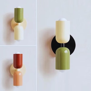 Peut inclure: Appliques murales modernes en trois couleurs : vert et crème, rouge et pêche, et vert et crème. Chaque applique présente un design cylindrique avec une base ronde et une ampoule en bas. Les appliques sont montées sur un mur blanc.