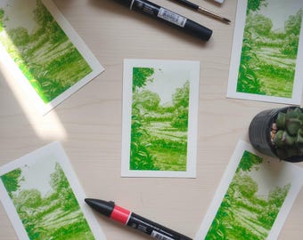Può includere: Diverse stampe artistiche di paesaggi verdi raffiguranti un sentiero attraverso una foresta. Le stampe sono accompagnate da materiali artistici, tra cui pennarelli neri e una piccola pianta grassa in un vaso nero, disposti su una superficie di legno chiaro.