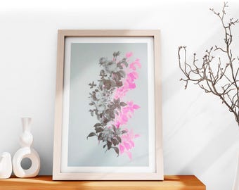 May include: A framed floral print featuring a vertical arrangement of flowers in shades of gray, pink, and white. The artwork is displayed in a light-colored wooden frame and rests on a wooden surface.