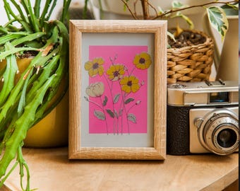 May include: A framed floral print featuring yellow and white flowers on a pink background. The artwork is displayed in a light-colored wooden frame. The image is set on a wooden surface, with plants and a vintage camera in the background.