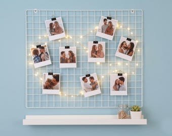 May include: A white grid photo display with several polaroid-style photos clipped to it. The photos feature couples in various poses. A string of warm white fairy lights is woven through the grid. A white shelf holds a jar and a small plant.
