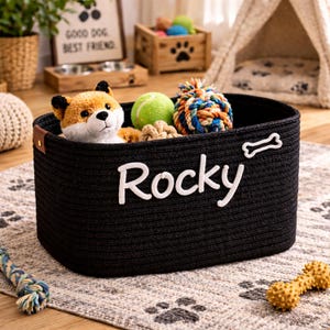 May include: A black woven rope pet toy basket with the name "Rocky" in white script. The basket contains a plush fox toy, a tennis ball, and other dog toys. A white bone-shaped accent is on the side.