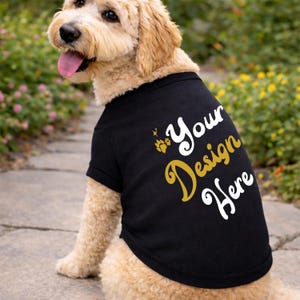 Puede incluir: Una camiseta negra para perros con el texto "Your Design Here" en dorado y blanco. La camiseta presenta una huella de pata y un diseño de estrella. El perro es de color marrón claro con la lengua rosa.