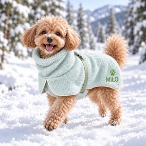 Peut inclure: Un manteau pour chien en polaire texturée vert clair avec un col montant et une sangle assortie. Le manteau présente une empreinte de patte brodée en vert et le nom MILO sur le côté. Le vêtement a un liseré clair sur les bords et une texture douce.