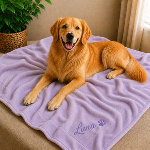 May include: A golden retriever dog lies on a lavender fleece blanket. The blanket has the name "Luna" embroidered in cursive, along with a paw print. The dog is smiling with its tongue out. A potted plant is in the background.