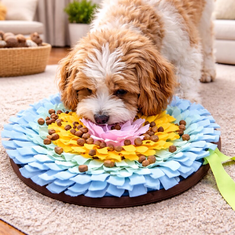 Op de afbeelding: Een hond snuffelt aan een kleurrijke, bloemvormige voermat voor huisdieren. De mat heeft lagen blauwe, gele en roze stoffen bloemblaadjes, met bruine brokjes verspreid. Een bruine rand omringt de mat.