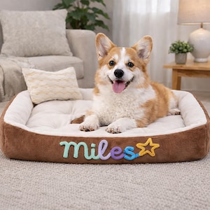 May include: A brown and white Corgi dog lying in a plush dog bed. The bed is brown with a cream-colored interior and the name "Miles" embroidered in colorful letters. A star is next to the name.
