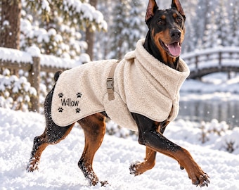 May include: A Doberman dog wearing a cream-colored, textured fleece coat with a high collar and a tan adjustable strap. The coat features the name Willow embroidered in black script, surrounded by four small black paw print icons on the side.
