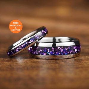 Può includere: Due anelli in metallo color argento con un intarsio centrale di frammenti di pietra iridescente viola e blu. Gli anelli presentano una finitura lucida e riflettente e un design a fascia arrotondata, mostrando una texture multicolore.
