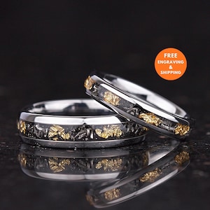 May include: Two polished silver-toned metal rings featuring a central inlay of black material embedded with small, irregular gold-colored flakes and dark metallic shavings. The rings have a smooth, reflective finish and a rounded band profile.