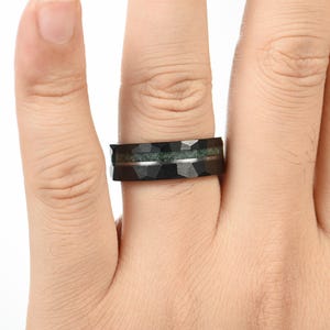 Puede incluir: Un anillo negro con una textura de superficie geom&eacute;trica facetada. La banda presenta una incrustaci&oacute;n horizontal de material texturizado de color verde musgo en el centro, separada de los bordes negros por una fina l&iacute;nea met&aacute;lica plateada.