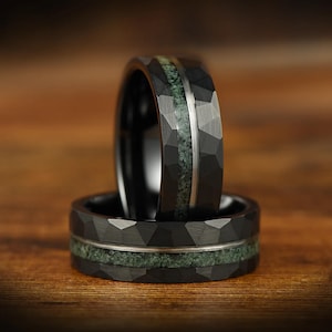 Puede incluir: Dos anillos negros con textura martillada y facetada. Cada anillo presenta una incrustaci&oacute;n horizontal central de material texturizado en color verde salvia, separado del metal negro por una fina l&iacute;nea plateada pulida en el contorno.