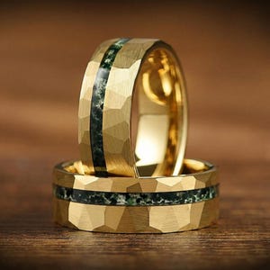 Op de afbeelding: Twee gouden ringen met een gehamerde textuur en een donkergroene stenen inleg. De ringen zijn op elkaar gestapeld, wat het ontwerp benadrukt. De achtergrond is warm hout. Trouwringen.