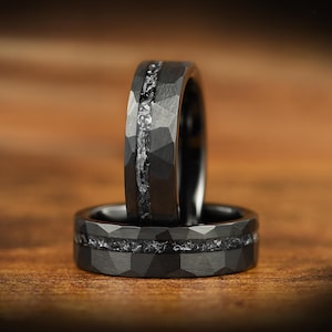 May include: Two dark gray rings with a hammered, faceted texture. Each band features a central horizontal inlay of crushed, shimmering metallic material that runs continuously around the circumference of the ring, creating a distinct contrast.
