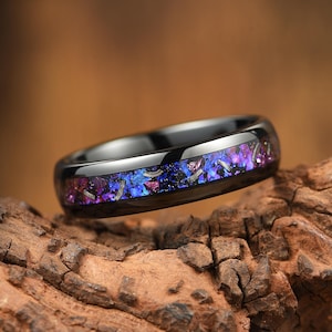 Peut inclure: Une bague noire polie avec une incrustation centrale composée de fragments violets, bleus et métalliques sombres. L'anneau présente une surface lisse et réfléchissante ainsi qu'un profil arrondi, révélant un motif moucheté multicolore.