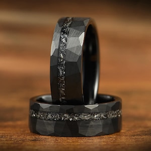 Könnte beinhalten: Ein schwarzer Ring mit einer facettierten, gehämmerten Außenfläche und einer mittigen Einlage aus texturierten, metallisch wirkenden Fragmenten. Das Band hat ein dunkles, mattes Finish mit geometrischen Facetten, die das Licht einfangen und einen Kontrast zur rauen, körnigen Textur der Einlage bilden.