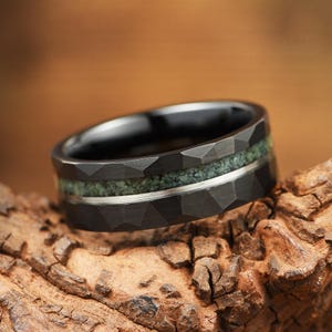 Puede incluir: Un anillo negro con una superficie exterior facetada que presenta una incrustaci&oacute;n central de material verde texturizado y una fina banda met&aacute;lica de tono plateado que recorre horizontalmente el centro de la incrustaci&oacute;n verde, creando contraste.