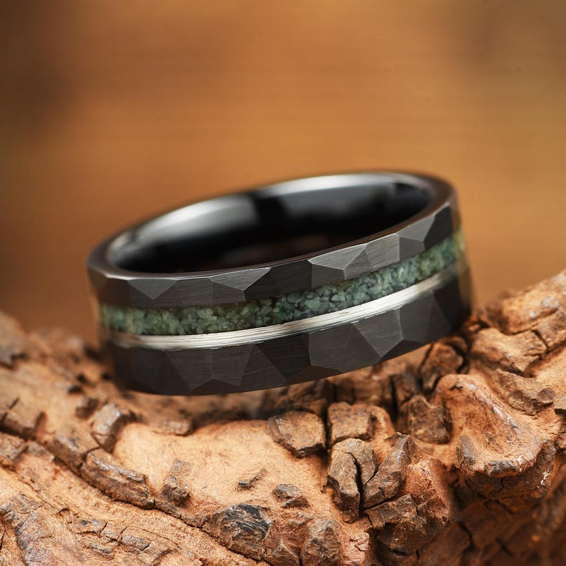 Puede incluir: Un anillo negro con una superficie exterior facetada que presenta una incrustaci&oacute;n central de material verde texturizado y una fina banda met&aacute;lica de tono plateado que recorre horizontalmente el centro de la incrustaci&oacute;n verde, creando contraste.