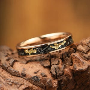 Könnte beinhalten: Ein Ring aus Roségold mit einer zentralen Einlage aus schwarzem Material, das mit metallischen Goldflocken gesprenkelt ist. Das Band hat eine polierte Roségold-Oberfläche an der Innenseite und den Kanten, die die dunkle, texturierte Einlage einrahmt.