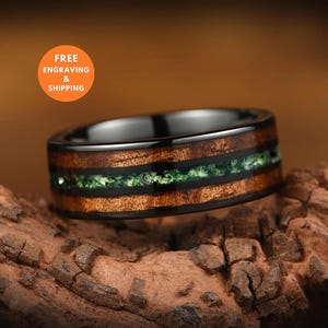 May include: A ring featuring a black metal band with a central inlay of brown wood and a thin, shimmering green crushed mineral stripe. The design includes two black metal borders framing the wood and green inlay, creating a layered appearance.
