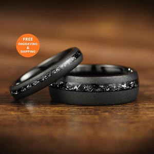 Könnte beinhalten: Zwei mattschwarze Ringe mit einer zentralen Einlage aus zerkleinertem Metallmaterial. Die Ringe haben eine texturierte, dunkle Oberfläche und ein glattes Innenband. Eine orangefarbene kreisförmige Grafik oben links zeigt den Text FREE ENGRAVING & SHIPPING.
