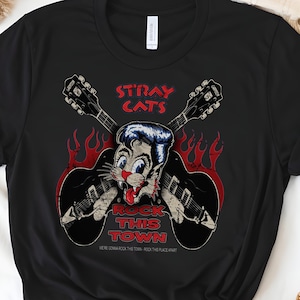 May include: Black t-shirt featuring a cartoon cat with blue hair and a red tongue, holding a guitar. The text "STRAY CATS" and "ROCK THIS TOWN" are in red. The shirt also has the text "WE'RE GONNA ROCK THIS TOWN - ROCK THIS PLACE APART".