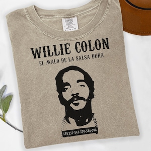 May include: A beige t-shirt featuring a black graphic of Willie Colón with the text "WILLIE COLON" and "EL MALO DE LA SALSA DURA". The shirt also has a phone number printed at the bottom. The shirt has a crew neck and short sleeves.
