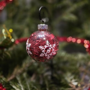 May include: A red Christmas ornament with a silver cap and a black hook. The clear glass ball is filled with red glitter and decorated with white snowflake designs. The ornament hangs in front of a green Christmas tree.