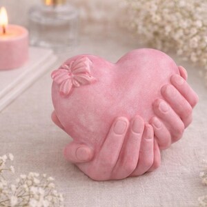 Può includere: Una scultura rosa a forma di cuore tenuta da due mani, con un dettaglio a farfalla. Una candela rosa accesa e fiori bianchi sullo sfondo. È un oggetto decorativo.